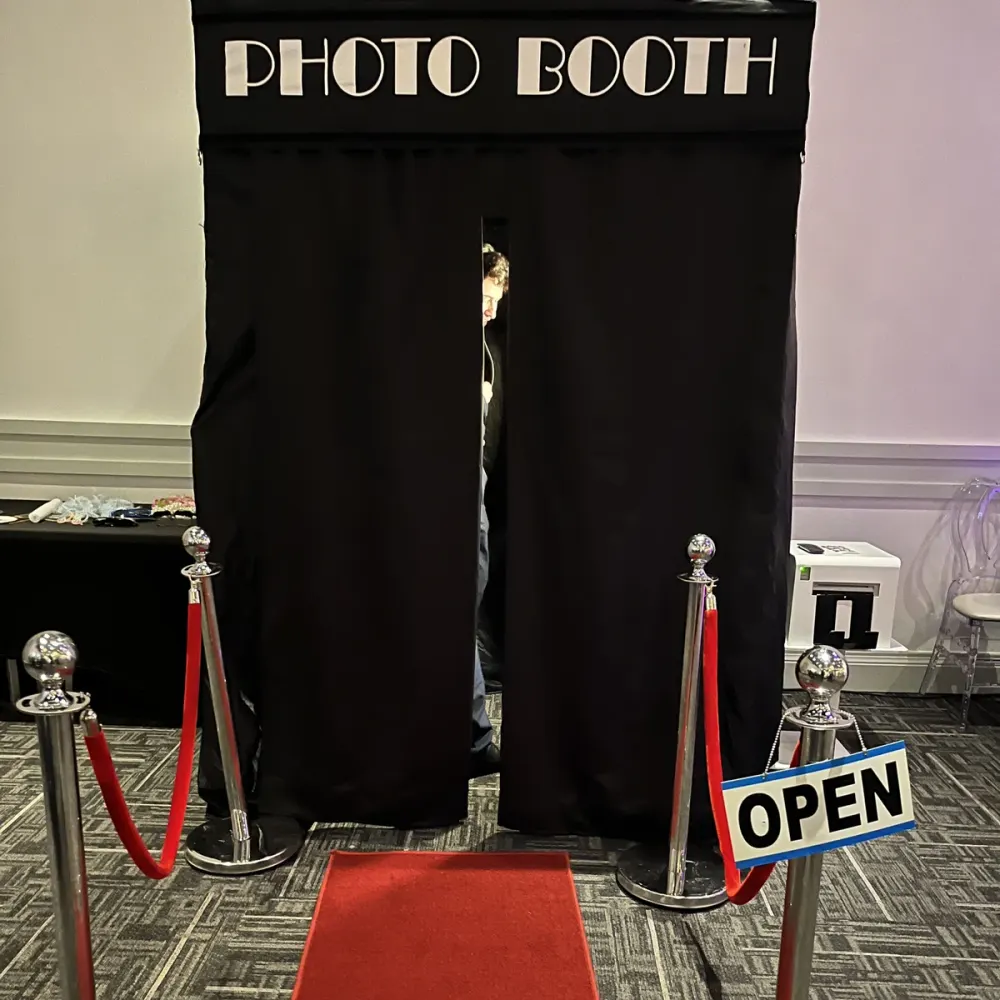 Enclosed private photo booth with black curtains, a red carpet, stanchions and an Open sign