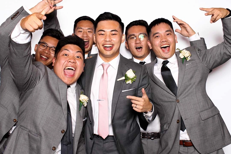 Groom and groomsmen in grey suits cheering in an open-air photo booth with a white backdrop: wedding photo booth rental in Burlington