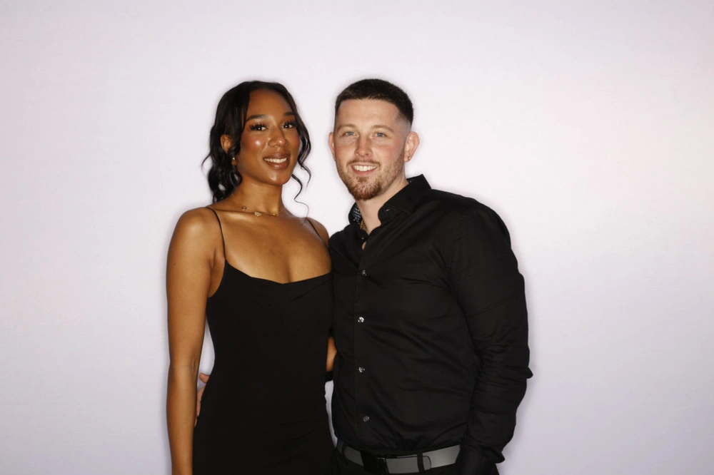 A couple dressed in black smiling against a white photo booth backdrop: photo booth rental in Hamilton