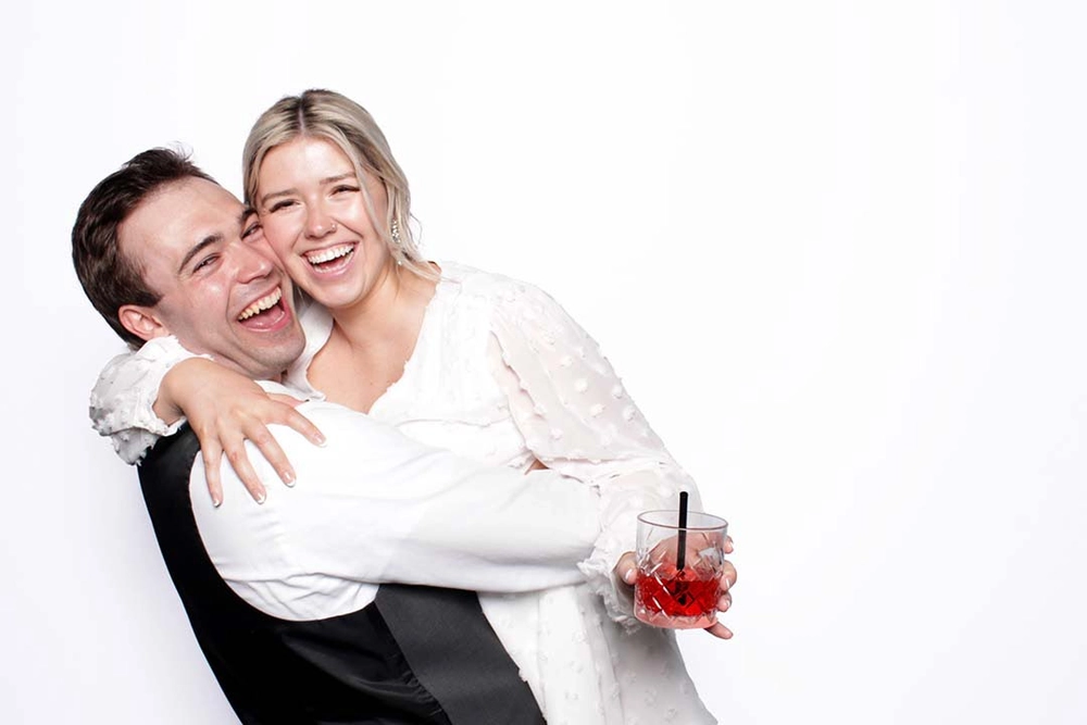 Couple laughing in a wedding photo booth against a white backdrop, one holding a cocktail: wedding photo booth rental in Hamilton