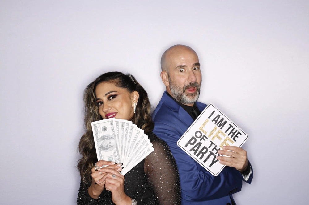 Two guests posing with a fan of prop banknotes and an I Am The Life Of The Party sign against a white backdrop: instant photo booth rental in Vaughan