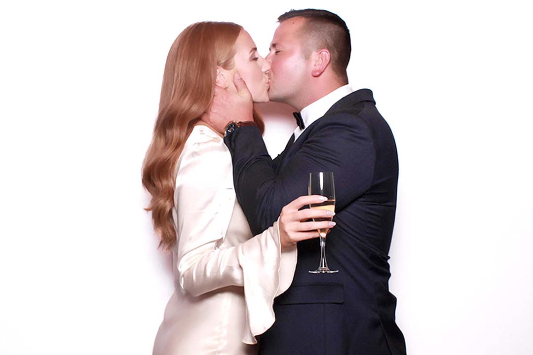 Elegant photo of a couple kissing in formal attire holding a champagne glass against a white backdrop, perfect for wedding photobooth rentals in Oakville.